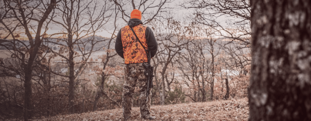 Hunter in orange vest and camouflage pants standing in a forested area holding a rifle