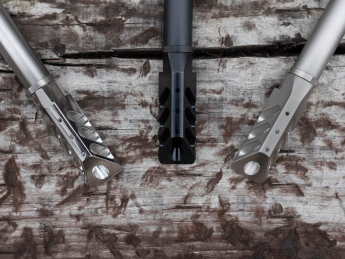 Three rifle muzzle brakes in different finishes displayed on a wooden surface.