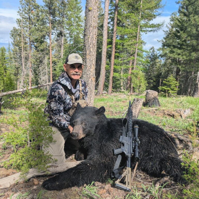 Man posing with a hunted black bear and a rifle in a forest clearing.