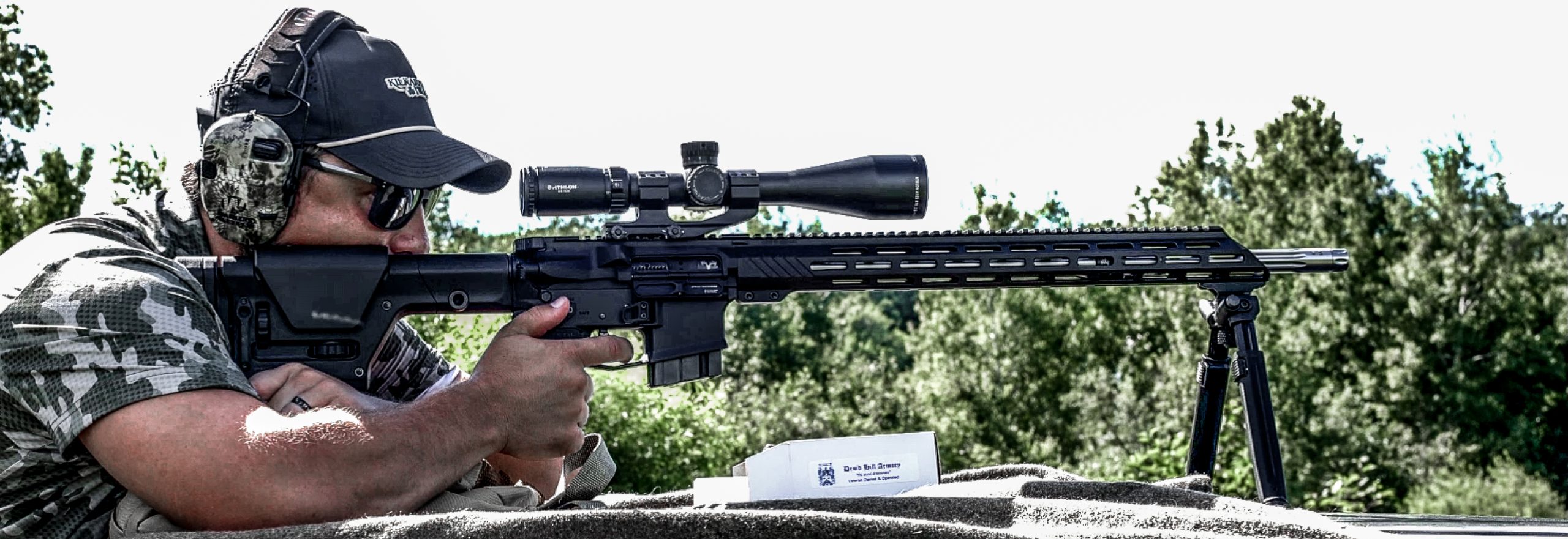 Person aiming a scoped rifle outdoors using a bipod wearing camo clothing and ear protection.