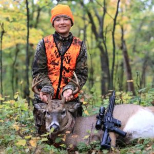 Hunter in orange vest posing with a deer and rifle in a forest setting.