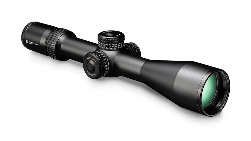 Black rifle scope with adjustment knobs and green lens on a white background.
