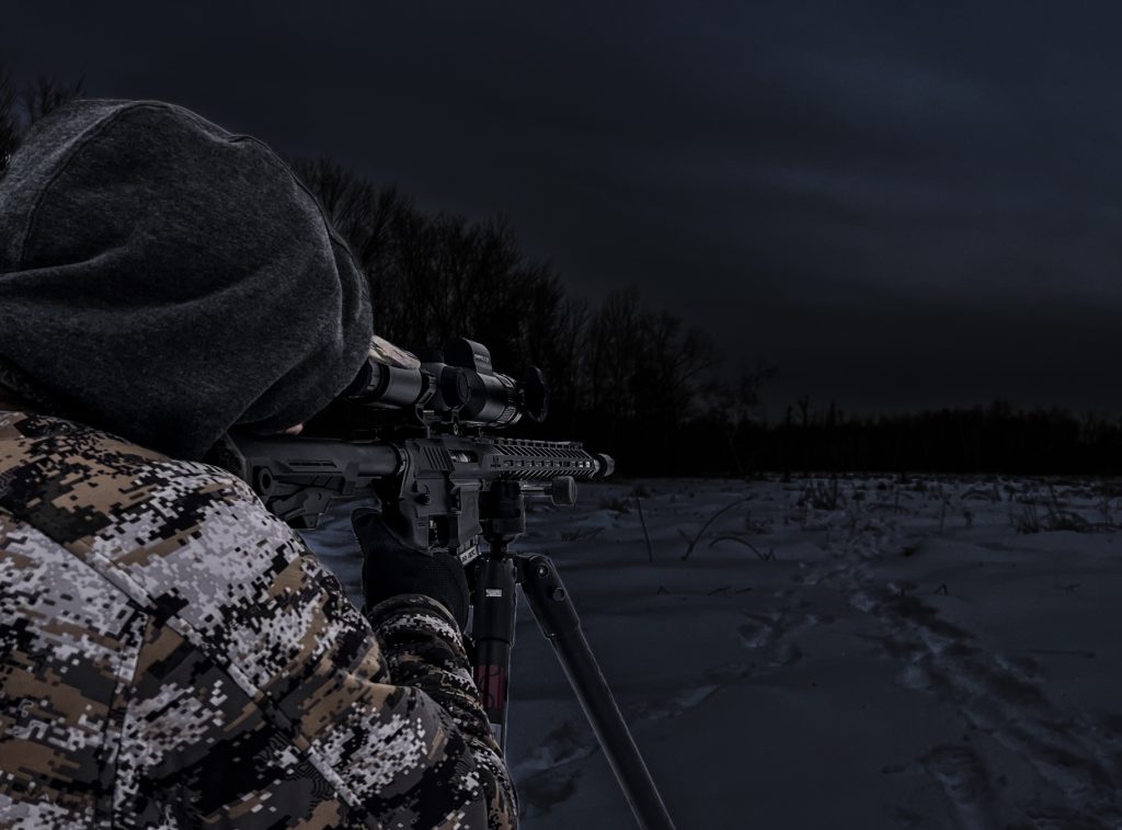 Person in camouflage aiming a rifle on a tripod in a snowy field at night.