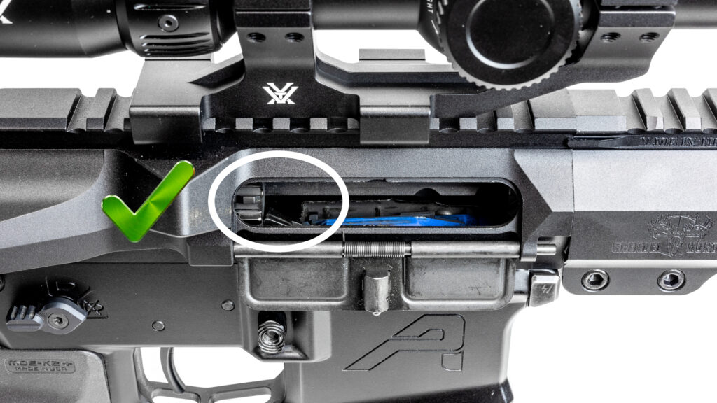 Close-up of an AR-15 ejection port with a green checkmark, demonstrating the correct bolt position where the bolt face is resting cleanly behind the bolt catch.