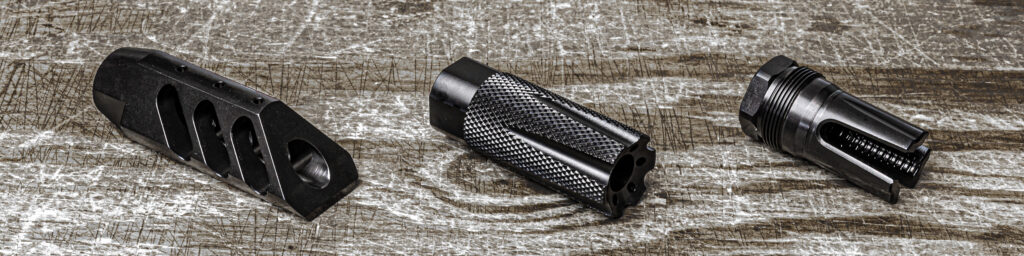 A product compilation photo displaying three distinct matte black AR-15 muzzle devices side-by-side on a rustic, weathered grey wood background. From left to right, the lineup showcases a multi-port precision muzzle brake, a knurled linear compensator with front gas ports, and a tactical open-prong flash hider with external suppressor attachment threads.