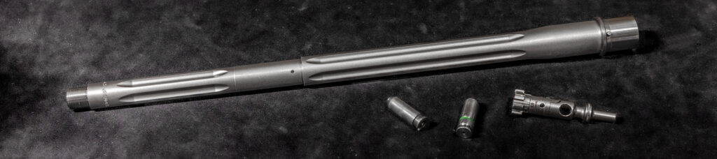 A close-up view displaying a custom rifle barrel with prominent straight fluting, threaded muzzle, and a matte, bead blasted stainless steel finish. Accompanying the barrel are its factory-matched precision bolt components, emphasizing a numbers-matched setup designed for optimal headspace and accuracy.